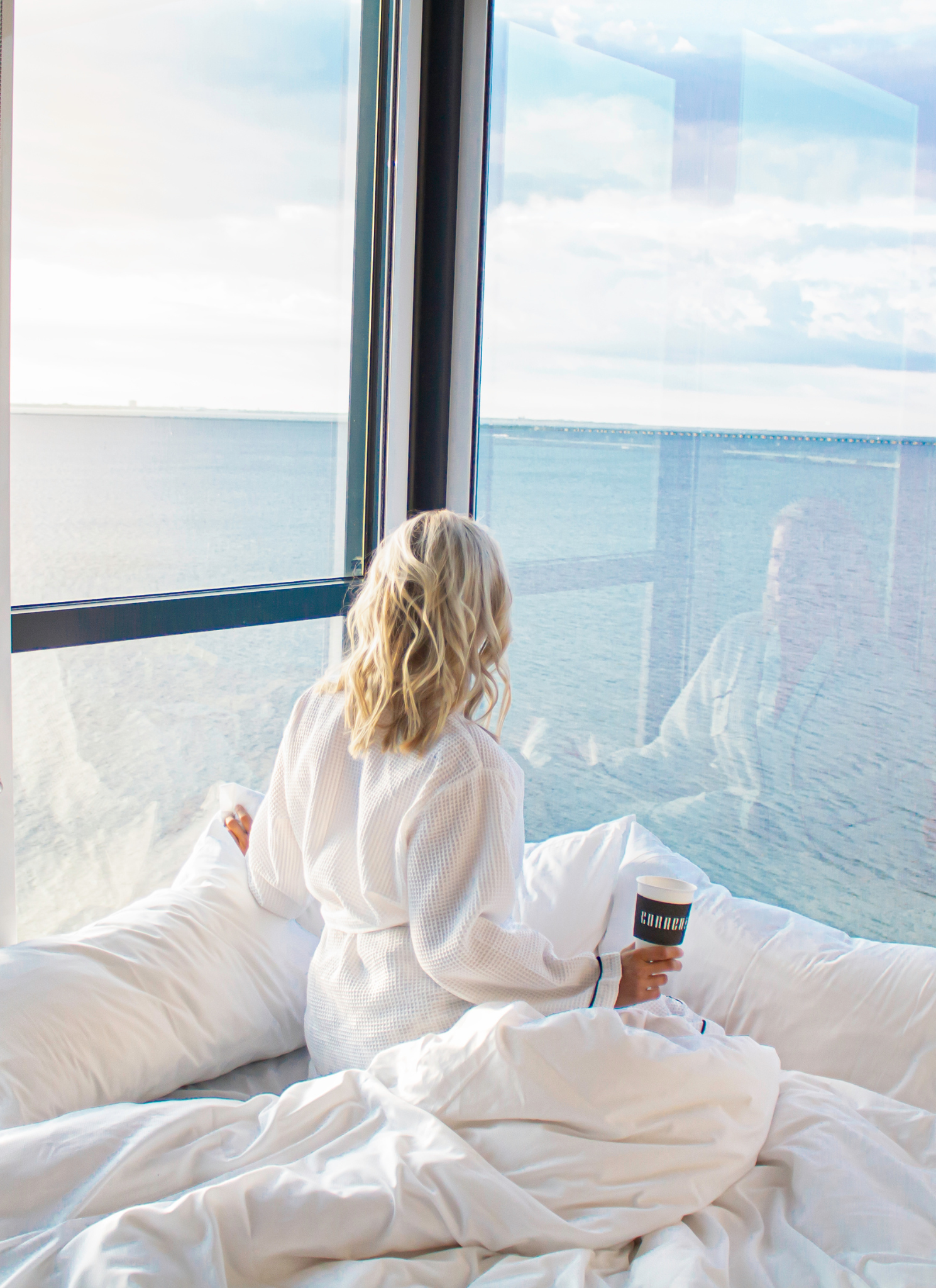 Travel Blogger, The Lovely Flamingo, Jill DiGioia wears a white robe looking out a 360 view of Tampa Bay at the Current Hotel Tampa