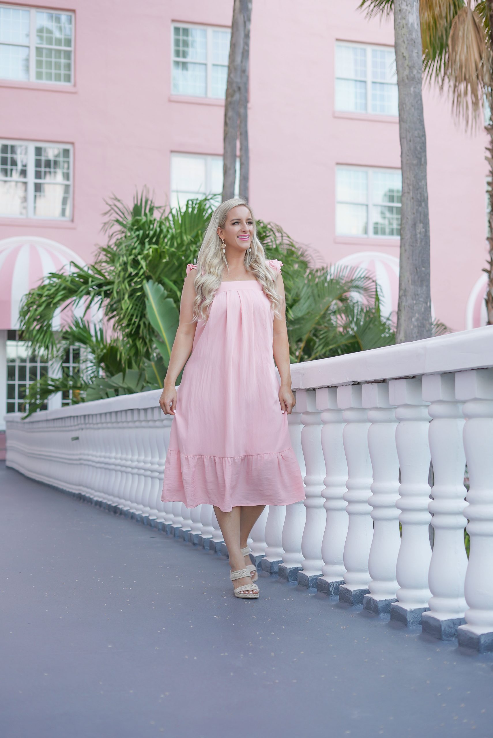 Florida Blogger, Jill DiGioia, The Lovely Flamingo wears gold shell earrings with pearl drop from her new beach earring collection with a pink midi beach dress standing in front of the Don Cesar on St. Pete Beach.