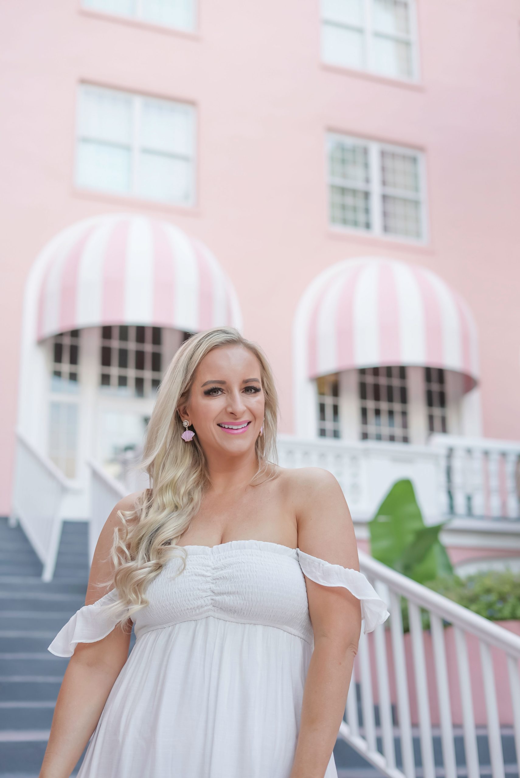 Florida Blogger, Jill DiGioia, The Lovely Flamingo wears pink shell earrings with pearl clusters from her new beach earring collection along with a white off the shoulder maxi dress standing in front of the Don Cesar on St. Pete Beach
